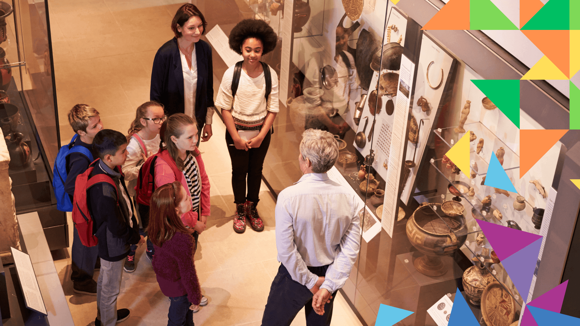 Image of group of children and teacher at museum, speaking with a member of staff. Grŵp o blant ac athro mewn amgueddfa yn siarad â aelod staff.